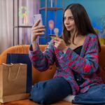Szczęśliwa shopaholic młoda kobieta siedząca z torbami na zakupy dokonując płatności online z kartą kredytową do domu