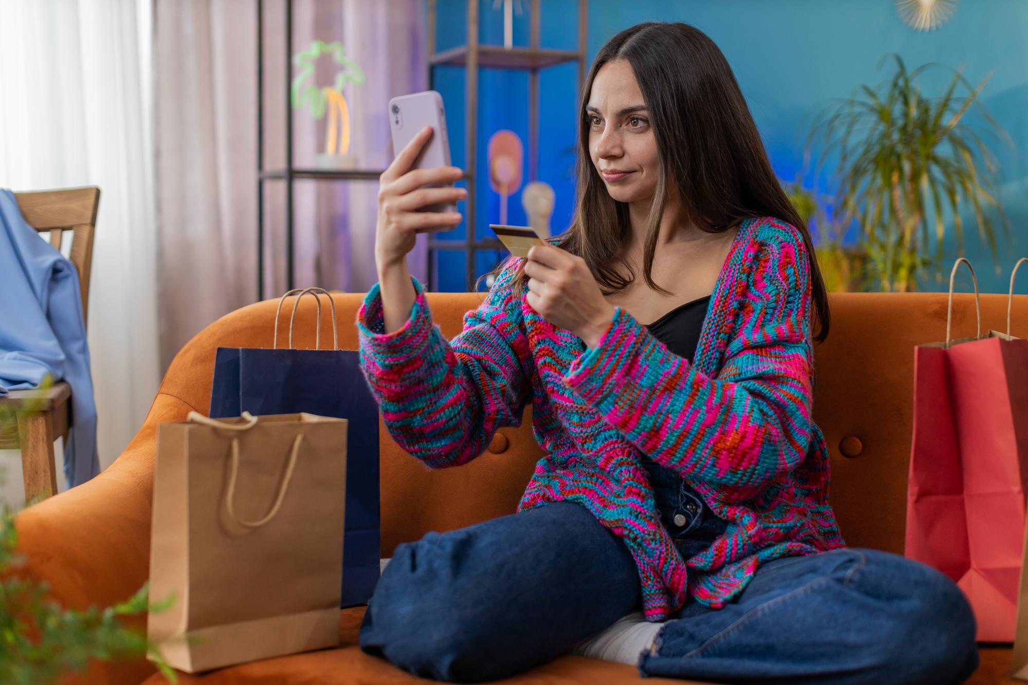 Szczęśliwa shopaholic młoda kobieta siedząca z torbami na zakupy dokonując płatności online z kartą kredytową do domu